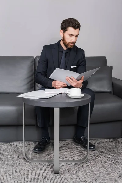 Hombre de negocios barbudo de traje mirando la carpeta cerca de la taza de café en la habitación de hotel - foto de stock