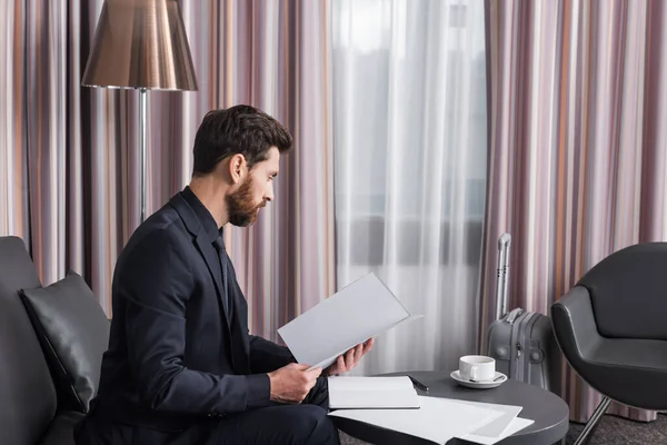 Vista lateral del hombre de negocios barbudo en traje mirando la carpeta con papeles cerca de la taza de café en la habitación de hotel - foto de stock