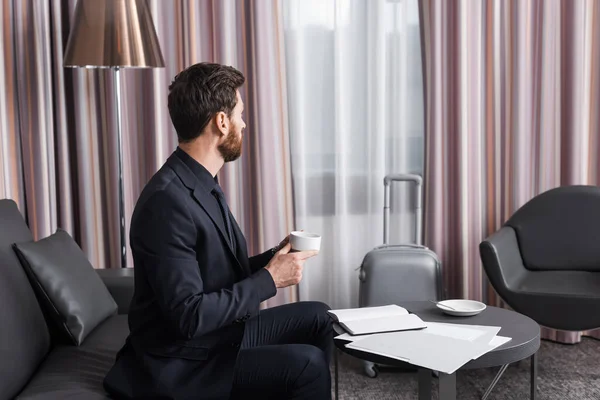 Bärtiger Mann im Anzug blickt auf Gepäck, während er eine Tasse Kaffee im Hotelzimmer hält — Stockfoto