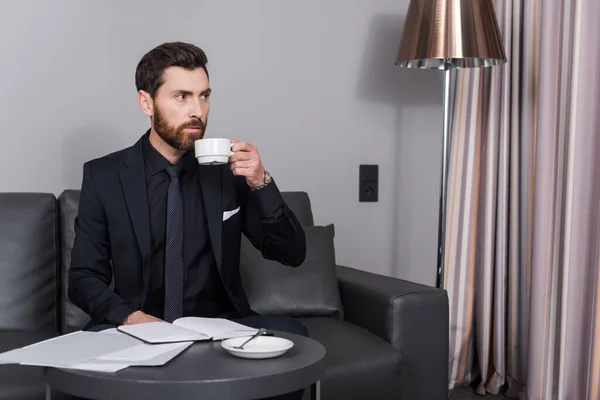 Uomo barbuto in giacca e cravatta in possesso di tazza di caffè vicino a documenti in camera d'albergo — Foto stock