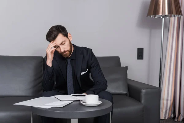 Müder Geschäftsmann im Anzug sitzt auf Couch neben Dokumenten und Tasse — Stockfoto