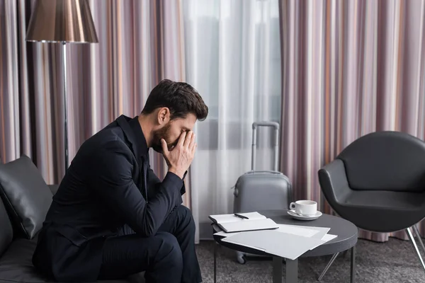 Seitenansicht eines müden Geschäftsmannes im Anzug, der sein Gesicht verdeckt und auf einer Ledercouch im Hotelzimmer sitzt — Stockfoto