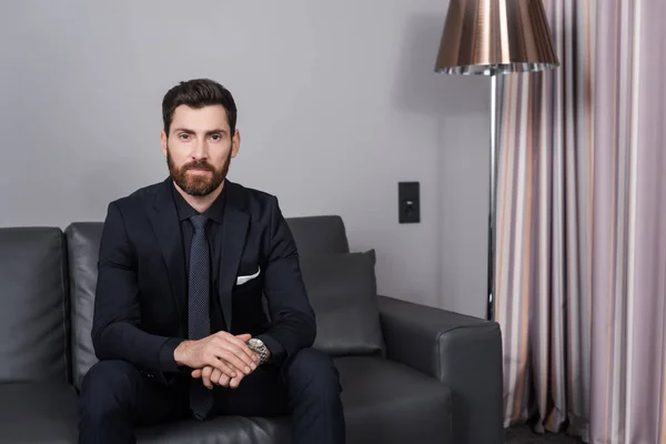 Bearded businessman in suit sitting on leather couch in hotel room — Stock Photo