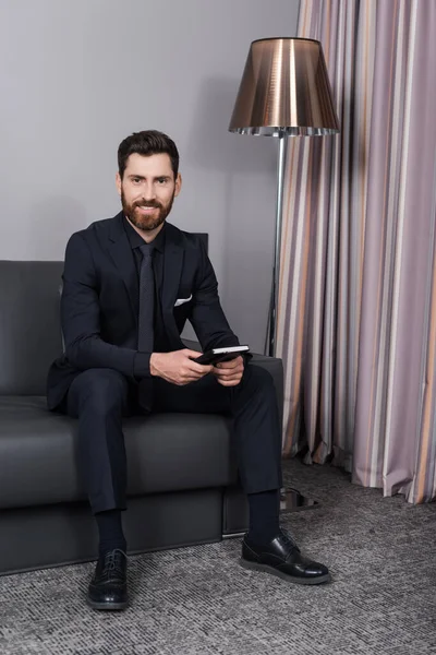 Happy man in suit holding notebook and sitting on leather couch in hotel room — Stock Photo