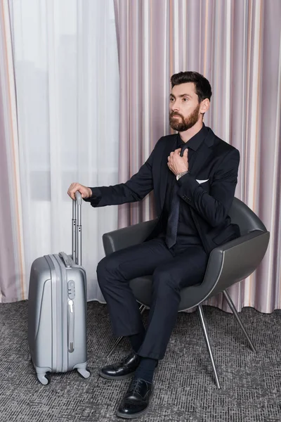 Hombre de negocios barbudo en traje ajustando corbata y sentado en sillón cerca de equipaje en la habitación de hotel - foto de stock