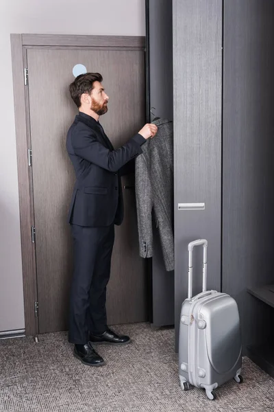 Full length of man in suit holding hanger with coat near closet and luggage in hotel room — Stock Photo