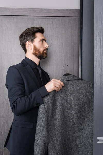 Side view of bearded man in suit holding hanger with coat near closet in hotel room — Stock Photo