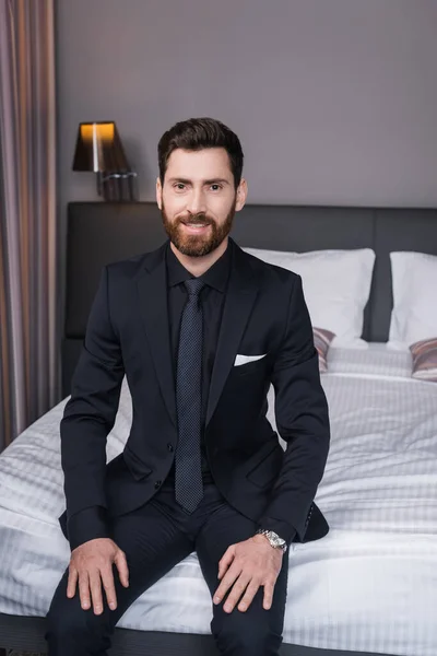 Bearded man in suit smiling while sitting on bed in hotel room — Stock Photo