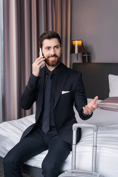 Щасливий чоловік у костюмі розмовляє по смартфону, сидячи на ліжку в готельному номері — стокове фото