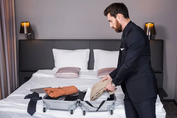 Side view of bearded man in suit unpacking suitcase in hotel room — Stock Photo