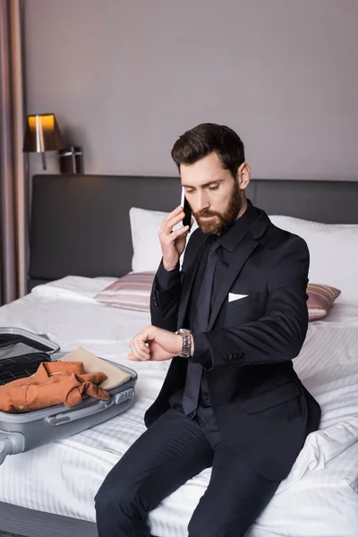 Hombre barbudo de traje hablando en el teléfono inteligente y el control de tiempo cerca de la ropa en el equipaje en la cama - foto de stock