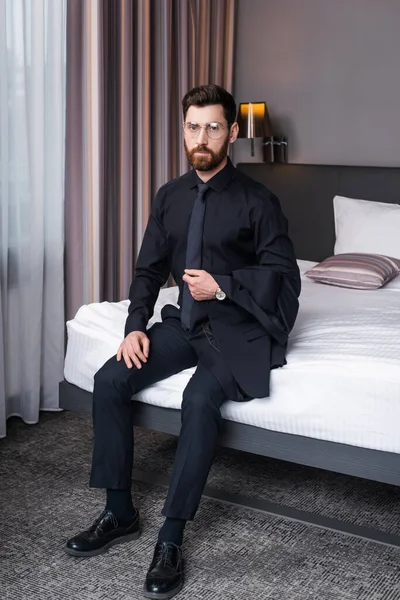 Bearded businessman in eyeglasses and suit sitting on bed in hotel room — Stock Photo