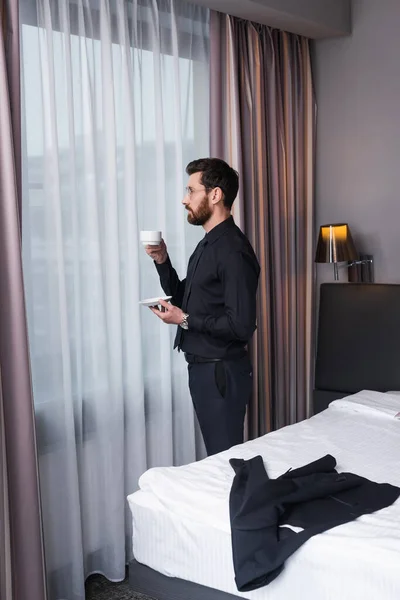 Homme barbu dans des lunettes et costume tenant tasse de café dans la chambre d'hôtel — Photo de stock