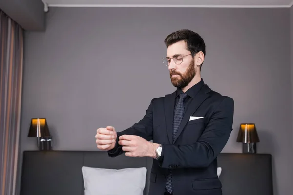 Bearded businessman in eyeglasses adjusting sleeve of shirt in hotel room — Stock Photo