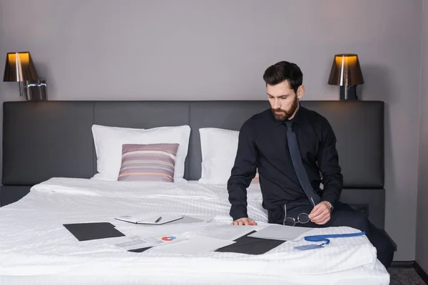 Bearded businessman holding eyeglasses and looking at documents on hotel bed — Stock Photo