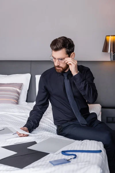 Bärtiger Mann im Anzug stellt Brille ein und schaut sich Dokumente auf Hotelbett an — Stockfoto