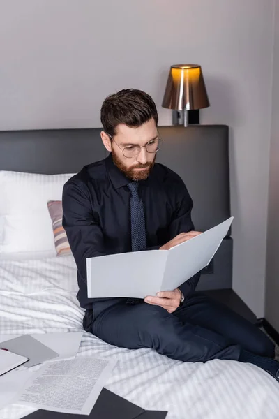 Бородатый мужчина в костюме и очках смотрит на папку и сидит на кровати отеля — стоковое фото