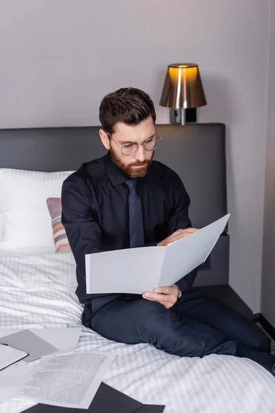 Bearded man in suit and eyeglasses looking at folder and sitting on hotel bed — Stock Photo