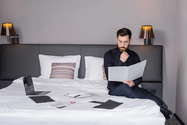 Hombre pensativo en traje y anteojos mirando a la carpeta y sentado cerca de la computadora portátil en la cama del hotel - foto de stock