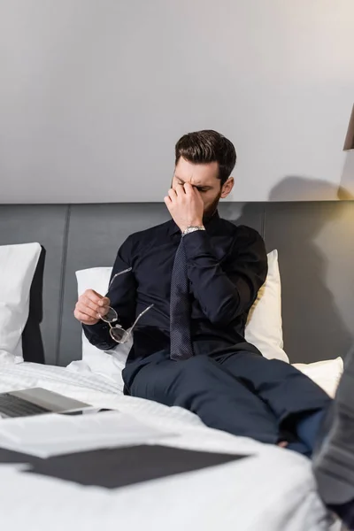 Exhausted bearded man in suit holding eyeglasses and sitting on hotel bed — Stock Photo
