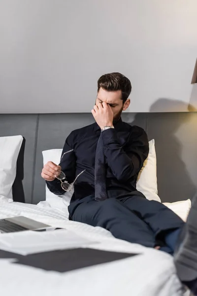 Hombre barbudo agotado en traje con gafas y sentado en la cama del hotel - foto de stock