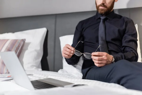 Cropped view of bearded man holding eyeglasses and sitting on hotel bed near laptop — Stock Photo