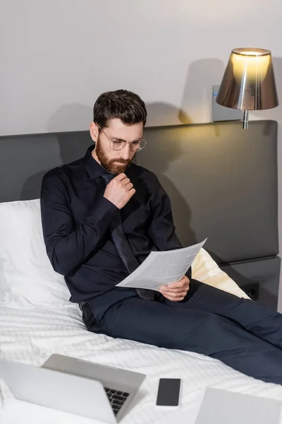 Homme barbu dans des lunettes de vue en regardant le document près de gadgets sur le lit de l'hôtel — Photo de stock