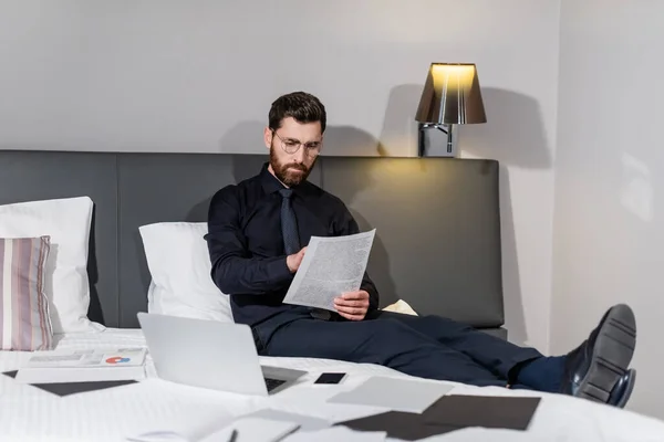 Hombre barbudo en anteojos mirando el documento cerca de aparatos e infografías en la cama del hotel - foto de stock