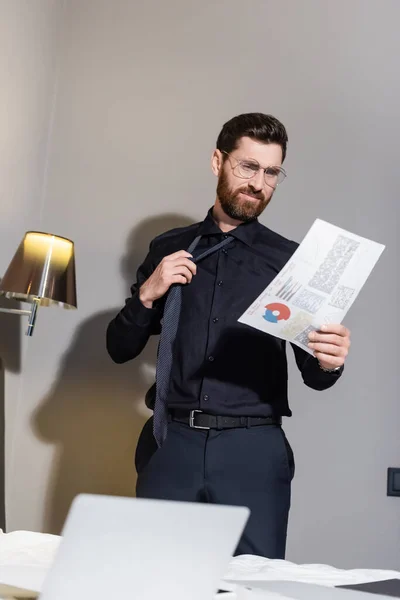 Homem barbudo em óculos ajustando gravata e olhando para infográficos no hotel — Fotografia de Stock