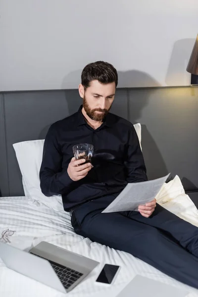 Uomo barbuto in possesso di un bicchiere di whisky e guardando il documento in hotel — Foto stock