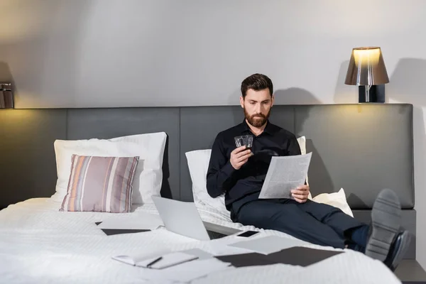 Uomo barbuto in possesso di un bicchiere di whisky e guardando il documento vicino gadget sul letto d'albergo — Foto stock