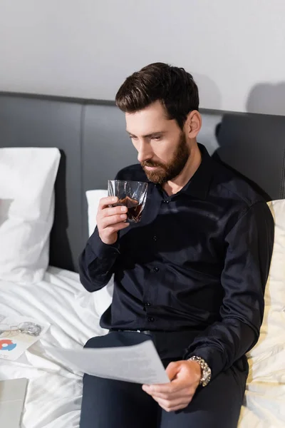 Bearded man holding glass of whiskey and looking at blurred document in hotel — Stock Photo