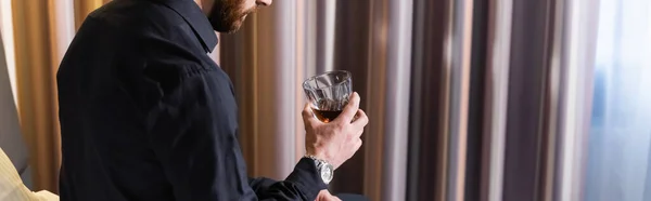 Vista ritagliata di uomo barbuto in possesso di un bicchiere di whisky e seduto nel letto d'albergo, banner — Foto stock