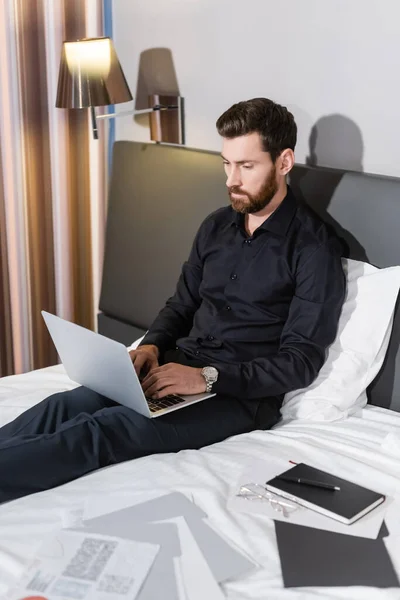 Hombre barbudo escribiendo en el ordenador portátil y sentado en la cama del hotel - foto de stock