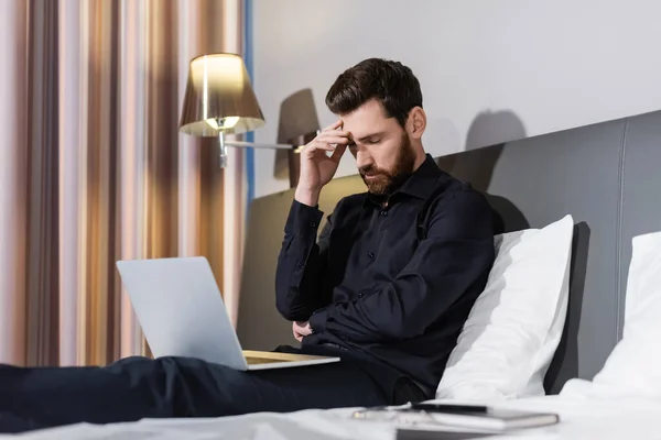 Уставший бородатый мужчина сидит с ноутбуком на кровати отеля — стоковое фото