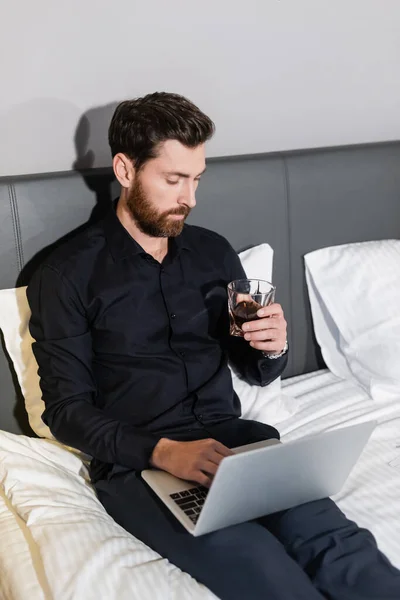 Barbudo hombre sosteniendo vaso de whisky y el uso de ordenador portátil en la cama del hotel - foto de stock