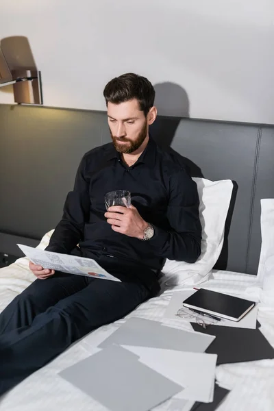 Homme barbu tenant un verre de whisky et assis dans le lit de l'hôtel près des infographies — Photo de stock