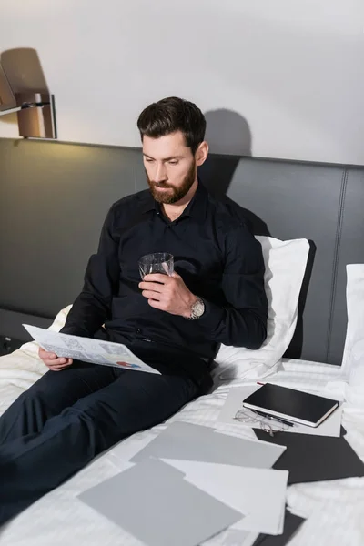 Bärtiger Mann mit einem Glas Whiskey im Hotelbett in der Nähe von Infografiken — Stockfoto