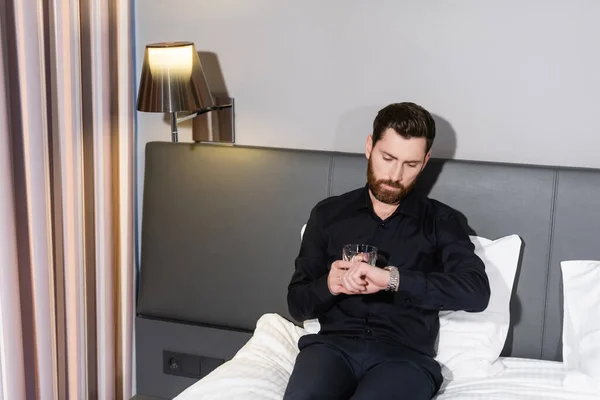 Homme barbu tenant un verre de brandy et de vérifier l'heure tout en étant assis dans le lit de l'hôtel — Photo de stock