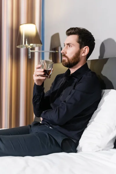 Hombre barbudo sosteniendo copa de brandy y sentado en la cama del hotel - foto de stock
