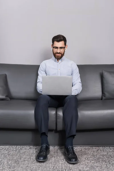 Happy man in eyeglasses and earphones using laptop while sitting on sofa in hotel — Stock Photo