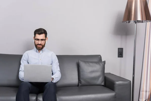 Cheerful man in eyeglasses and earphones using laptop while sitting on sofa in hotel — Stock Photo