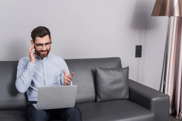 Hombre feliz en gafas ajustando los auriculares y el uso de la computadora portátil mientras está sentado en el sofá en el hotel - foto de stock