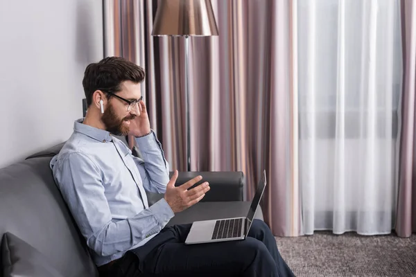 Side view of happy man in eyeglasses having video call while sitting on sofa in hotel — Stock Photo