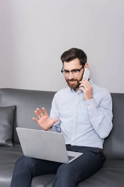 Улыбающийся мужчина в очках, разговаривающий по телефону, сидя с ноутбуком в отеле — стоковое фото