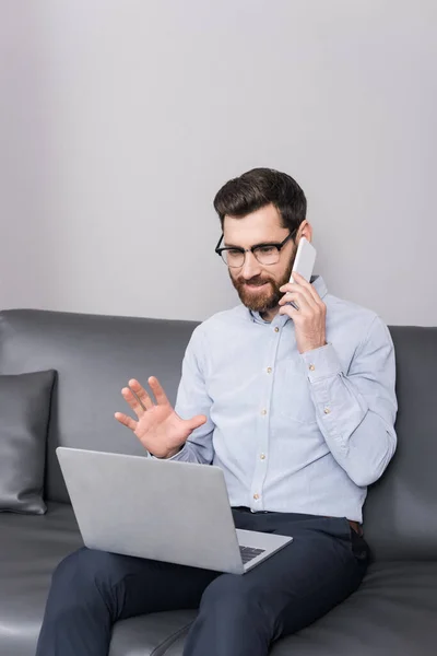 Uomo sorridente in occhiali avendo telefonata mentre seduto con il computer portatile in hotel — Foto stock