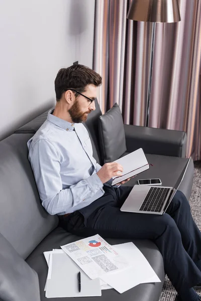 Bearded man in eyeglasses looking at notebook near devices on couch — Stock Photo