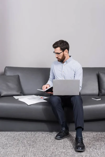 Bearded man in eyeglasses holding notebook near devices on couch — Stock Photo