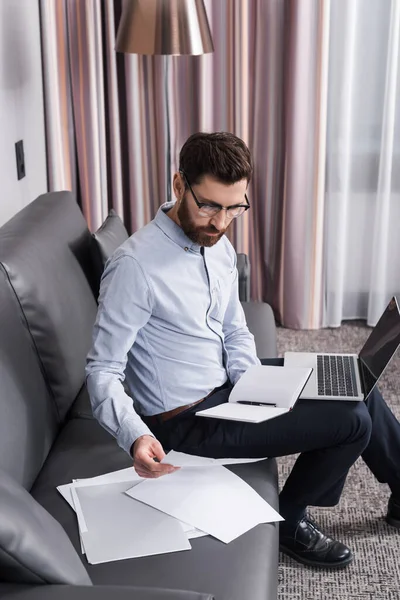 Hombre barbudo en gafas con documento y portátil cerca de la computadora portátil con pantalla en blanco - foto de stock