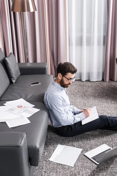 Hombre barbudo en anteojos sentado en la alfombra y la escritura en el cuaderno cerca de la computadora portátil y sofá - foto de stock